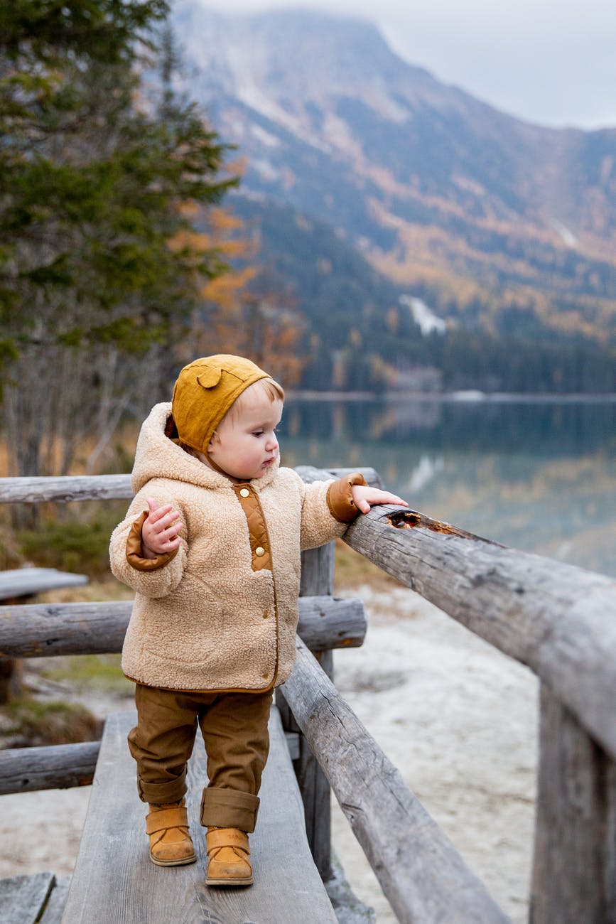 child in brown jacket standing on wooden fence near lake